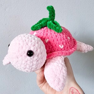 May include: A pink crocheted turtle with a strawberry shell and green leaves. The turtle has black eyes and a pink body.