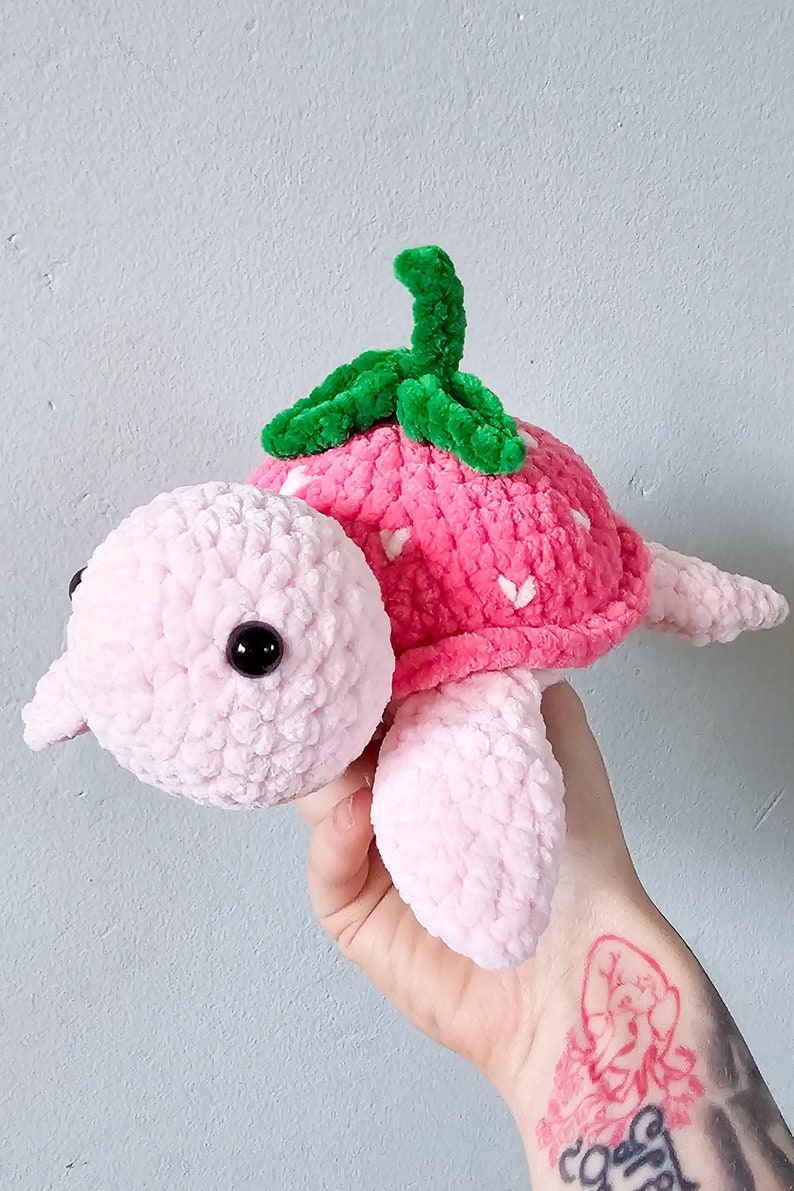 May include: A pink crocheted turtle with a strawberry shell and green leaves. The turtle has black eyes and a pink body.