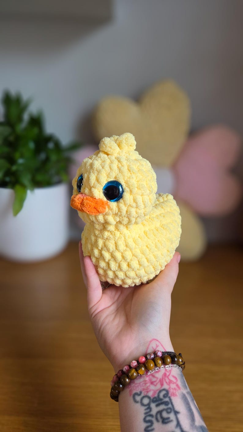 May include: A handmade, yellow crochet duck with large blue eyes and an orange beak. The plush toy is soft and textured, suitable for a child's room or as a decorative item. The duck is held in a hand.