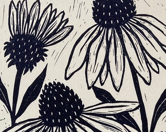 May include: Black and white linocut print of coneflowers and foliage. The artwork features three large coneflowers with prominent petals and dark centers, surrounded by various leaves and stems. The print has a textured, handmade appearance.