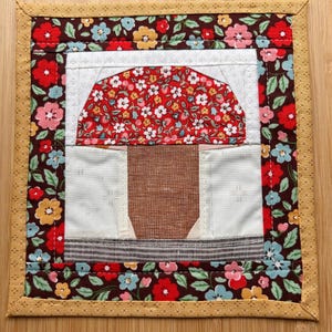 May include: A square fabric quilt block featuring a central mushroom motif with a red floral cap and a brown textured stem. The mushroom is set against a white background, framed by a dark brown border with a colorful floral pattern and a tan edge.