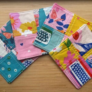 May include: A set of three square fabric coasters featuring a patchwork design with various colorful patterns including butterflies, flowers, typewriters, and ice cream cones. The coasters are quilted with visible stitching and have rounded corners.