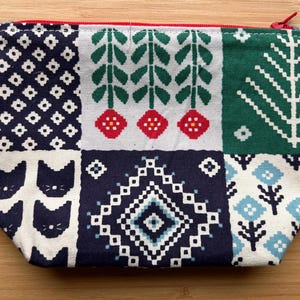 May include: A rectangular fabric zipper pouch featuring a patchwork design with various patterns including dark blue cat faces, white floral motifs, green pine trees, red flowers, and a central dark blue diamond shape with white geometric borders.