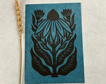 May include: A teal-colored card with a black floral linocut print featuring a central flower and surrounding leaves. A single stalk of wheat is positioned to the left of the card.
