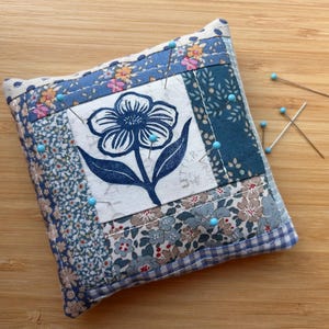 May include: A square fabric pincushion featuring a central white panel with a dark blue floral illustration. The surrounding border consists of various blue, white, and tan floral and gingham fabric patches. Several light blue pins are inserted.
