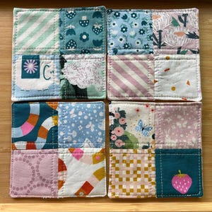 May include: Four square fabric coasters, each featuring a patchwork design of four distinct cotton squares stitched together. The patterns include floral, striped, strawberry, and animal motifs in a palette of teal, pink, blue, white, and orange.