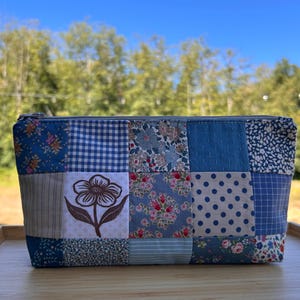May include: A rectangular zippered pouch featuring a patchwork design of various blue and white fabrics. The patterns include gingham, polka dots, small floral prints, and a single square panel displaying a dark brown line drawing of a flower.