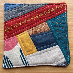 May include: A square fabric coaster featuring a patchwork design of various textile scraps in red, blue, mustard yellow, pink, and white. The piece includes decorative yellow embroidery stitching across the red section and orange running stitches.