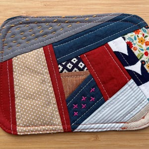 May include: A rectangular quilted fabric coaster featuring a patchwork design of various textiles including red, navy blue, beige polka dot, and floral patterns. The piece is accented with decorative yellow and pink hand-stitched embroidery details.