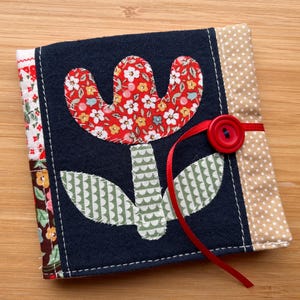 May include: A square fabric notebook cover featuring a dark blue center panel with a red floral applique flower and green patterned leaves. The cover includes side panels with white floral and beige polka dot patterns, secured by a red ribbon and button.