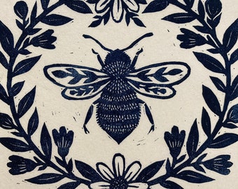 Puede incluir: Un grabado en linóleo que presenta una abeja detallada en el centro, rodeada por una corona de hojas, flores y capullos. El diseño está realizado en tinta azul oscuro sobre un fondo crema, creando un contraste llamativo. Las alas y el cuerpo de la abeja están intrincadamente detallados.
