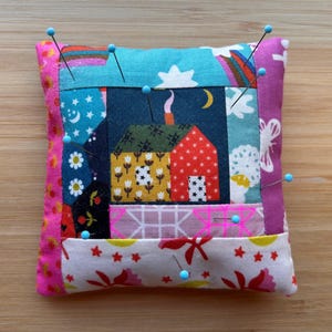 May include: A square fabric pincushion featuring a patchwork design with a central house illustration, surrounded by colorful patterns including stars, flowers, and butterflies. Several straight pins with light blue heads are inserted.