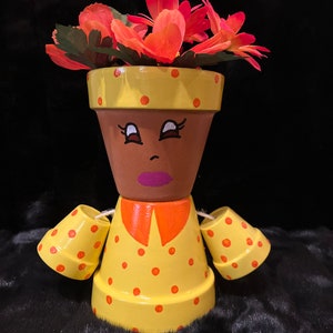 May include: A hand-painted terracotta planter shaped like a person wearing a yellow dress with orange polka dots and a red flower crown. The planter has a brown face with black eyes, a brown nose, and a pink mouth.
