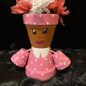May include: A pink and white polka dot planter with a painted face and white flowers on top. The planter is shaped like a person with arms and a dress.