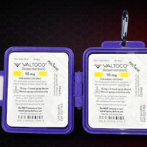 May include: Two purple plastic cases containing Valtoco diazepam nasal spray, each labeled 10 mg for nasal use only. The cases feature white informational labels with black text, a peel here tab, and a metal carabiner attached to the top of one case.