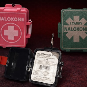 May include: A pink case labeled NALOXONE with a white cross, a dark green case labeled I CARRY NALOXONE with a star symbol, and an open black case containing a 4 mg Naloxone nasal spray unit with a barcode and expiration date of 2025-MR.