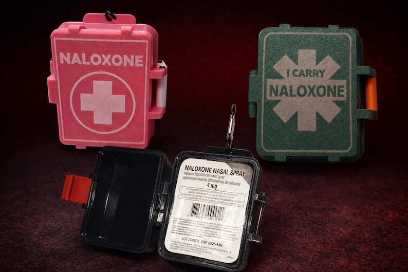 May include: Three rectangular plastic cases for naloxone nasal spray. One is pink with a white cross and the text NALOXONE. One is green with a star of life symbol and the text I CARRY NALOXONE. One is open, black, and contains a 4 mg nasal spray label.