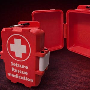 May include: A bright red, rectangular, hard-shell plastic case with a white cross symbol and the text "Seizure Rescue medication" printed on the front. The case is shown in two positions: one closed and one open, revealing an empty interior.