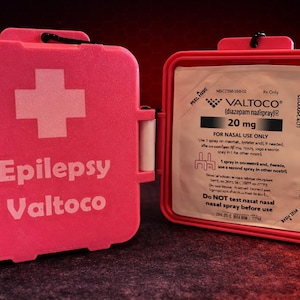 May include: A bright pink rectangular medical case with a white cross and the text Epilepsy Valtoco on the front. The case is open, revealing a white foil packet labeled Valtoco diazepam nasal spray 20 mg for nasal use only inside.