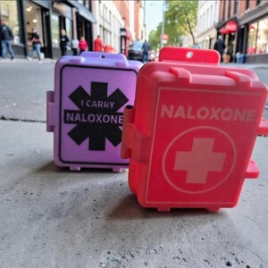 May include: Two rectangular plastic cases, one lavender and one red, designed for carrying naloxone. The lavender case features a black star of life symbol with the text I CARRY NALOXONE. The red case displays a white cross symbol and the word NALOXONE.