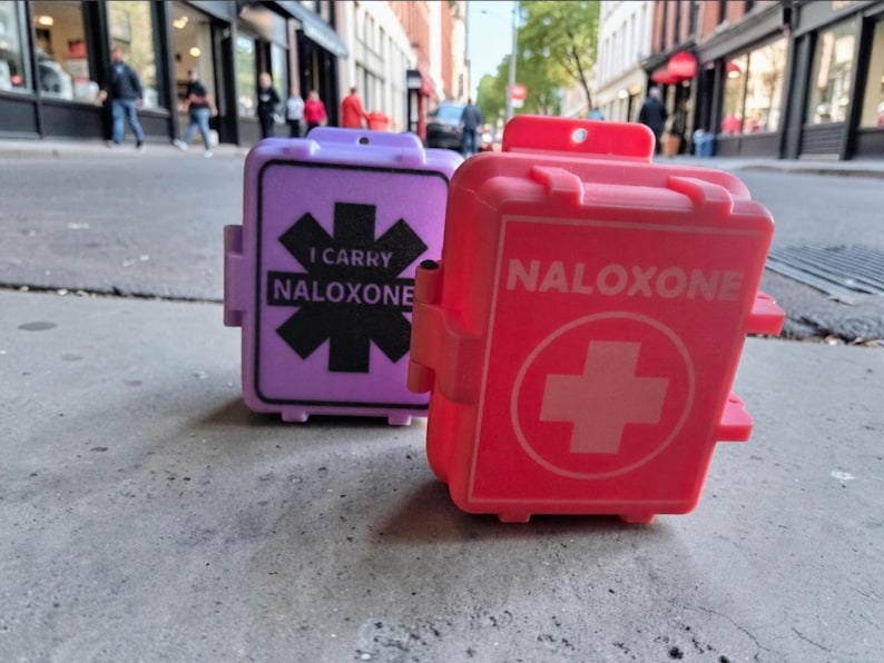 May include: Two rectangular plastic cases, one lavender and one red, designed for carrying naloxone. The lavender case features a black star of life symbol with the text I CARRY NALOXONE. The red case displays a white cross symbol and the word NALOXONE.