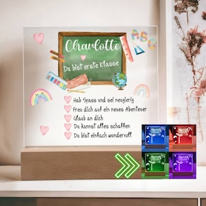 Op de afbeelding: Een gepersonaliseerd verlicht bord met een schoolbordachtergrond en de naam "Charlotte" in roze geschreven. Het bord bevat ook de tekst "Du bist erste Klasse" en een regenboog, een aardbol en schoolbenodigdheden. Het bord wordt verlicht in verschillende kleuren, waaronder rood, blauw en groen.