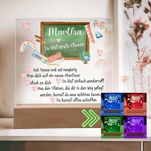 Op de afbeelding: Een gepersonaliseerd verlicht bord met een schoolbordachtergrond en de naam "Mantha" erop geschreven. Het bord bevat ook de tekst "Du bist erste Klasse" en een kleurrijke illustratie van schoolbenodigdheden. Het bord wordt verlicht in verschillende kleuren, waaronder blauw, rood, groen en paars.
