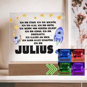 Op de afbeelding: Een verlicht bord met een raket- en astronautenontwerp. Het bord zegt "Ich bin stark, ich bin wichtig. Ich bin klug, ich bin mutig. Ich werde von Herzen geliebt. Ich bin genug, ich bin einzigartig. Ich glaube an mich. Ich kann alles schaffen. Ich bin Julius." Het bord is verlicht in blauw, rood, groen en paars.