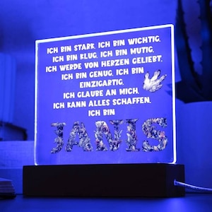 Op de afbeelding: Een blauw getinte LED-nachtlamp. Het licht toont de naam "JANIS" in een bloemenontwerp en Duitse tekst. Het licht staat op een donkere houten basis en heeft een wit snoer.