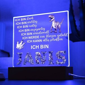 Op de afbeelding: Een blauw verlicht bord met een dinosaurus afbeelding en de tekst "ICH BIN stark, ICH BIN wichtig, ICH BIN mutig, ICH BIN einzigartig, ICH BIN wundervoll, ICH WERDE von Herzen geliebt, ICH KANN alles schaffen, ICH BIN JANIS".