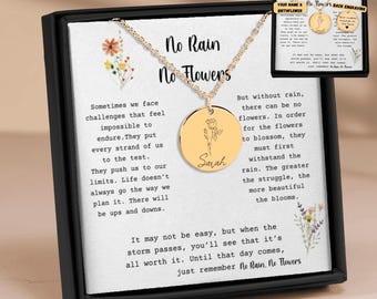 May include: A gold necklace with a round pendant featuring a delicate flower design and the name "Sarah" engraved on it. The necklace comes in a black box with a white card that reads "No Rain No Flowers" and a quote about overcoming challenges.