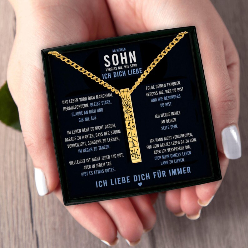 a person's hand holding a black box with a gold chain necklace inside. The box contains text in German, which appears to be a description or information about the necklace.