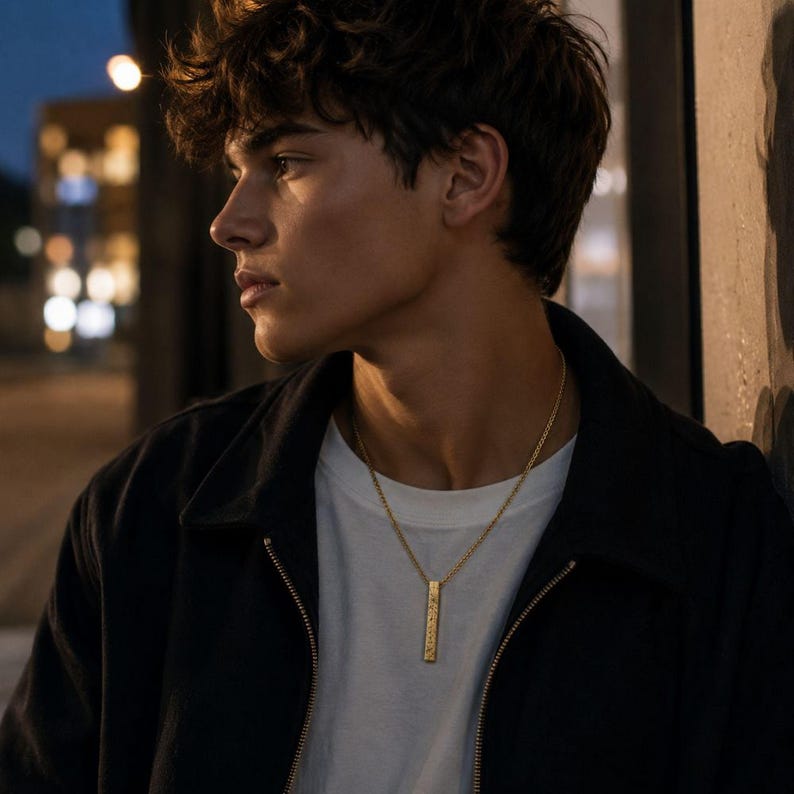 A young man with curly hair wearing a black jacket and a gold necklace stands in a dimly lit urban setting, gazing off to the side.