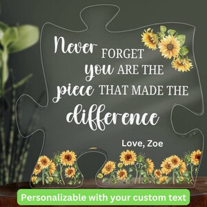May include: A clear acrylic puzzle piece shaped like a jigsaw piece with the text "Never FORGET you ARE THE piece THAT MADE THE difference Love, Zoe" and a sunflower design. The text is white and the sunflowers are yellow with green leaves.