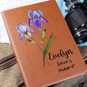 Puede incluir: Un diario de cuero marrón con una ilustración de acuarela de iris morados. El diario está personalizado con el nombre "Evelyn", el mes de "enero" y la palabra "snowdrop".