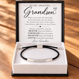 Puede incluir: Una pulsera de cordón trenzado negro con una barra de metal plateado que muestra el texto Your Text Here, presentada en una caja negra con una tarjeta de mensaje dirigida a un nieto, incluyendo la firma Love, Grandma.