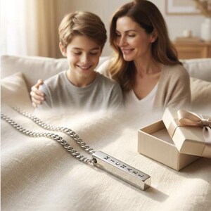 A mother and son are sitting together on a couch, smiling and looking at a necklace with a pendant that has the name "Michael" engraved on it.
