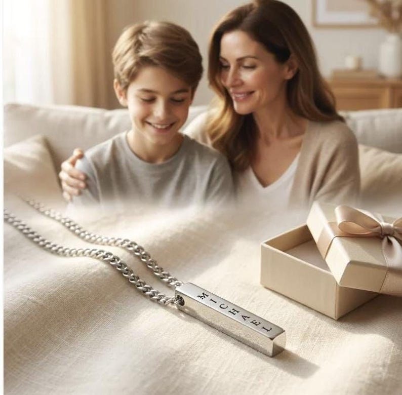 A mother and son are sitting together on a couch, smiling and looking at a necklace with a pendant that has the name "Michael" engraved on it.