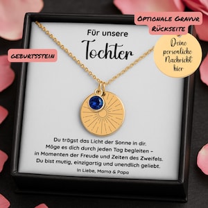 Op de afbeelding: Een goudkleurige ketting met een hanger met een blauwe edelsteen en een zonnestraalontwerp. De ketting wordt gepresenteerd in een zwarte doos met Duitse tekst, waaronder "Für unsere Tochter".