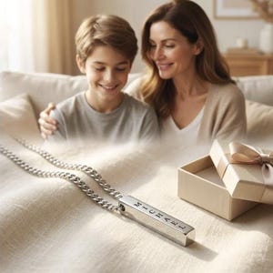 A mother and son are sitting together on a couch, smiling and looking at a necklace with a pendant that has the name "Michael" engraved on it.