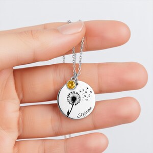 A hand holding a silver necklace with a round pendant featuring a dandelion design and a yellow gemstone.