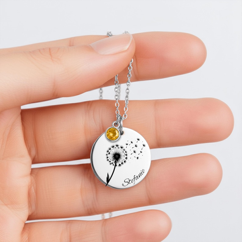 A hand holding a silver necklace with a round pendant featuring a dandelion design and a yellow gemstone.