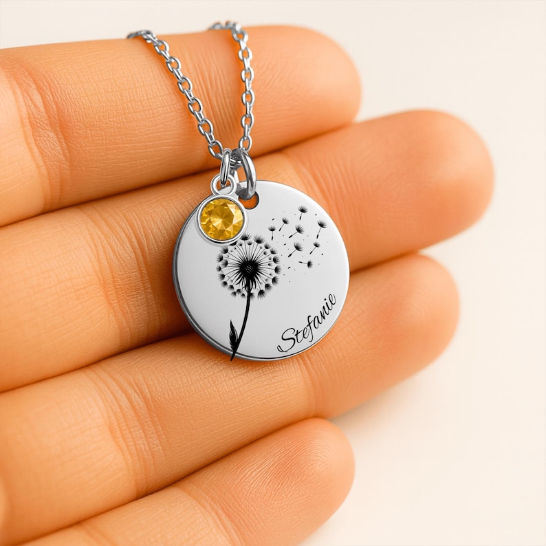 A hand holding a silver necklace with a round pendant featuring a dandelion design and a yellow gemstone.