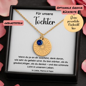 Op de afbeelding: Gouden ketting met een ronde hanger met een blauwe edelsteen en een gegraveerd zonneradiatie-ontwerp. De ketting wordt gepresenteerd in een zwarte doos met Duitse tekst, waaronder "Für unsere Tochter" en "Geburtsstein".