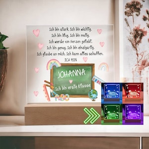 Op de afbeelding: Een gepersonaliseerd verlicht bord met een schoolbordachtergrond en de tekst "ICH BIN JOHANNA Ich bin eerste klasse" in het Duits. Het bord is omringd door roze harten, een regenboog en schoolbenodigdheden. Het bord is verkrijgbaar in vier kleuren: blauw, rood, groen en paars.