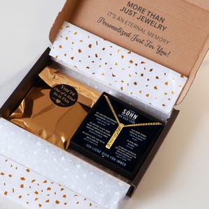 a gift box with a gold-colored item inside, and a card with text on it. The box is open, revealing a gold-colored item and a black card with text on it.