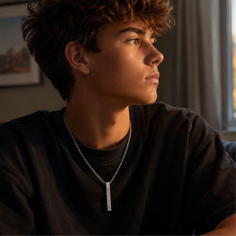 A young man with curly hair wearing a black shirt and a necklace is looking off to the side, with a thoughtful expression.