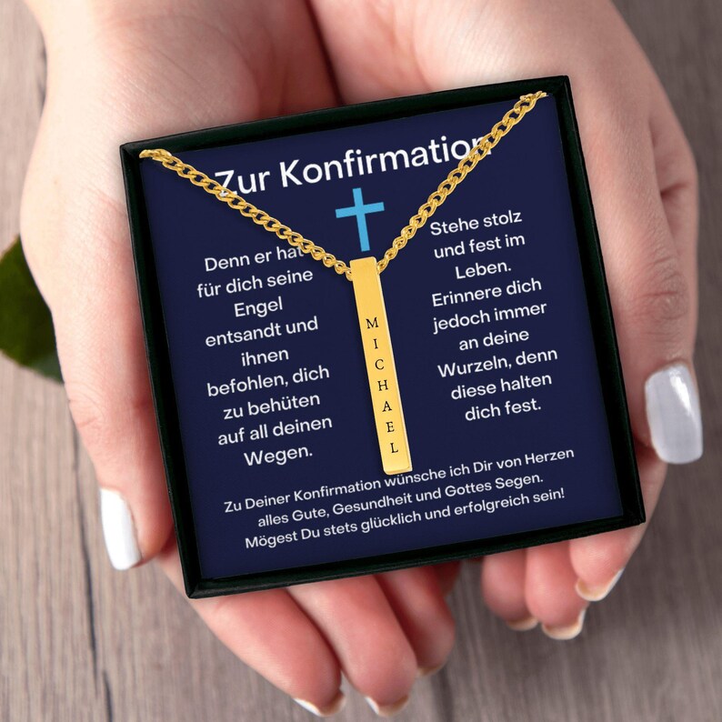 a person's hands holding a necklace with a gold chain and a blue background. The necklace has a gold cross on it. The text on the necklace reads "Zur Confirmation" and "Michael".