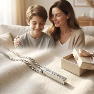 A mother and son are sitting together on a couch, smiling and looking at a necklace with a pendant that has the name "Michael" engraved on it.