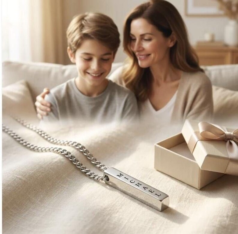 A mother and son are sitting together on a couch, smiling and looking at a necklace with a pendant that has the name "Michael" engraved on it.